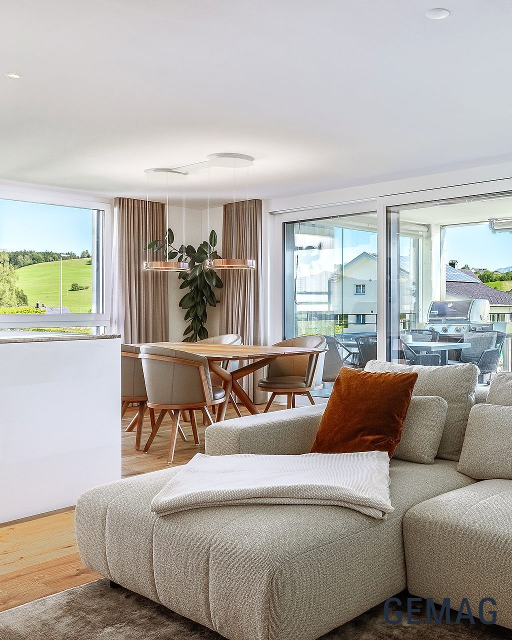 Offener Wohn-Essbereich mit grossem Sofa, Esstisch und bodentiefen Fenstern mit Blick auf Balkon und Hügellandschaft.