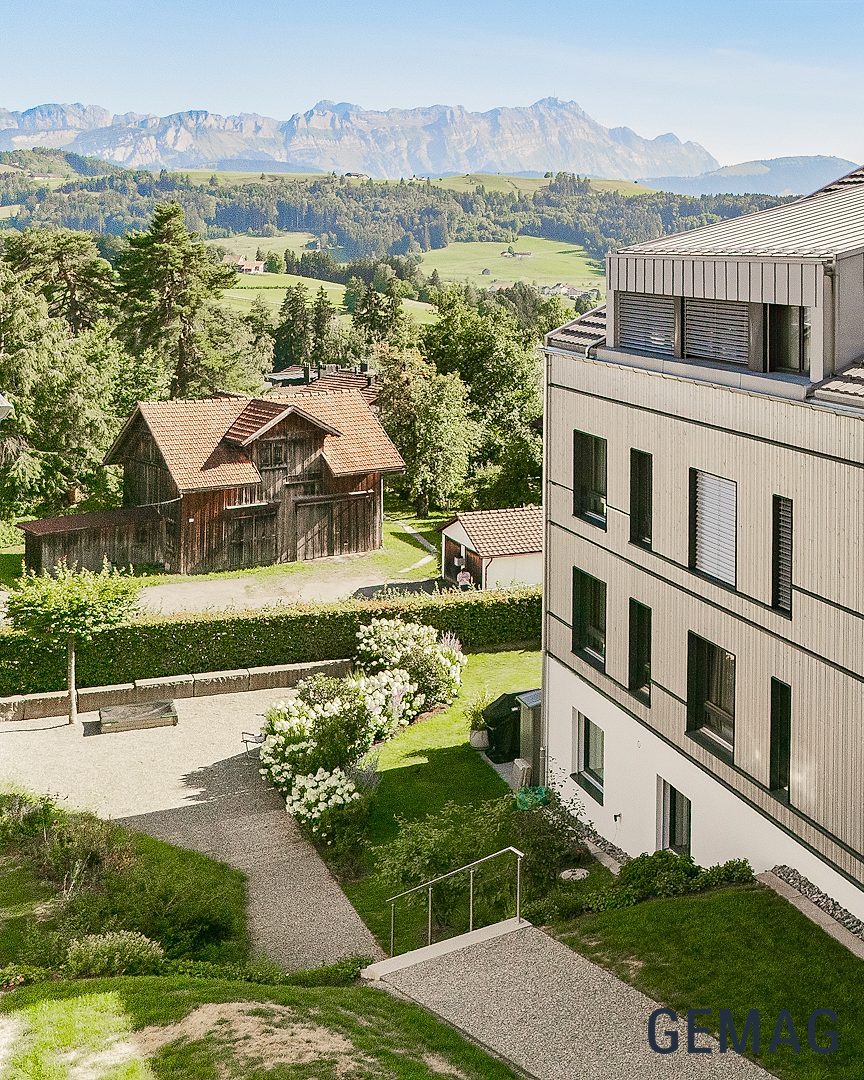 Mehrfamilienhaus mit heller Holzverkleidung und Satteldach, Schrägluftaufnahme mit Grünumgebung und Bergen im Hintergrund.