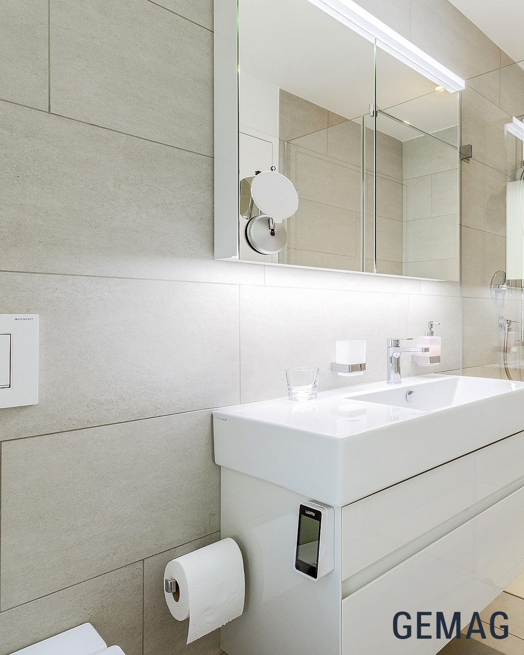 Badezimmer mit Waschtischunterschrank, Spiegelschrank und bodenebener Glasdusche, hellgraue Wandfliesen.