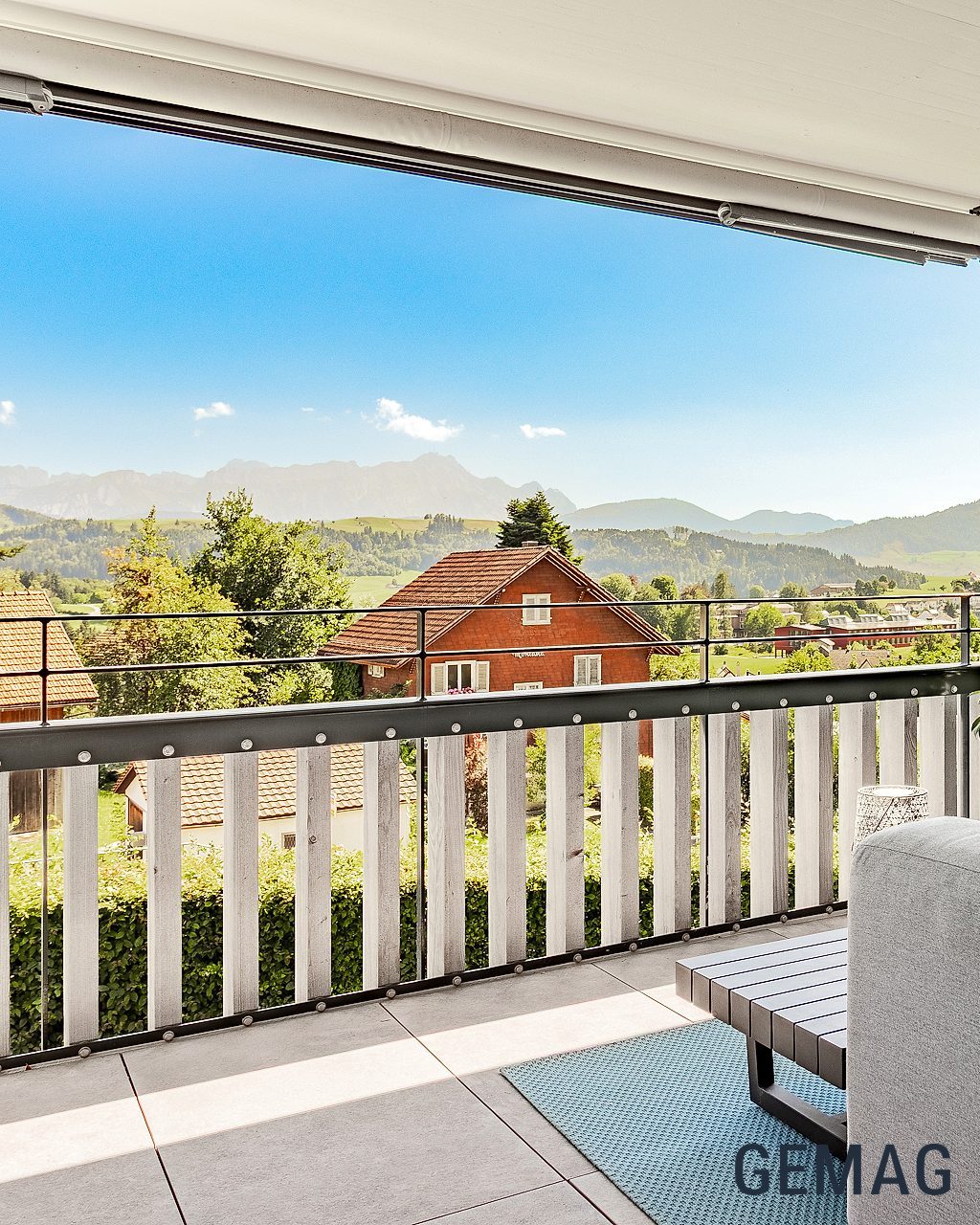 Überdachter Balkon mit Sitzbereich und weitem Blick auf Berge und Ortschaft bei sonnigem Wetter.