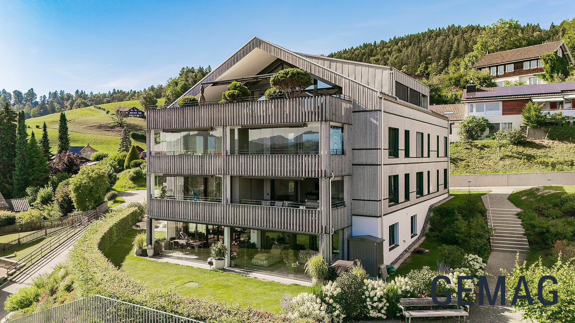 Mehrgeschossiges Wohngebäude mit Holzlamellenfassade und verglasten Balkonen, umgeben von Grünflächen und Hügeln, Drohnenaufnahme.