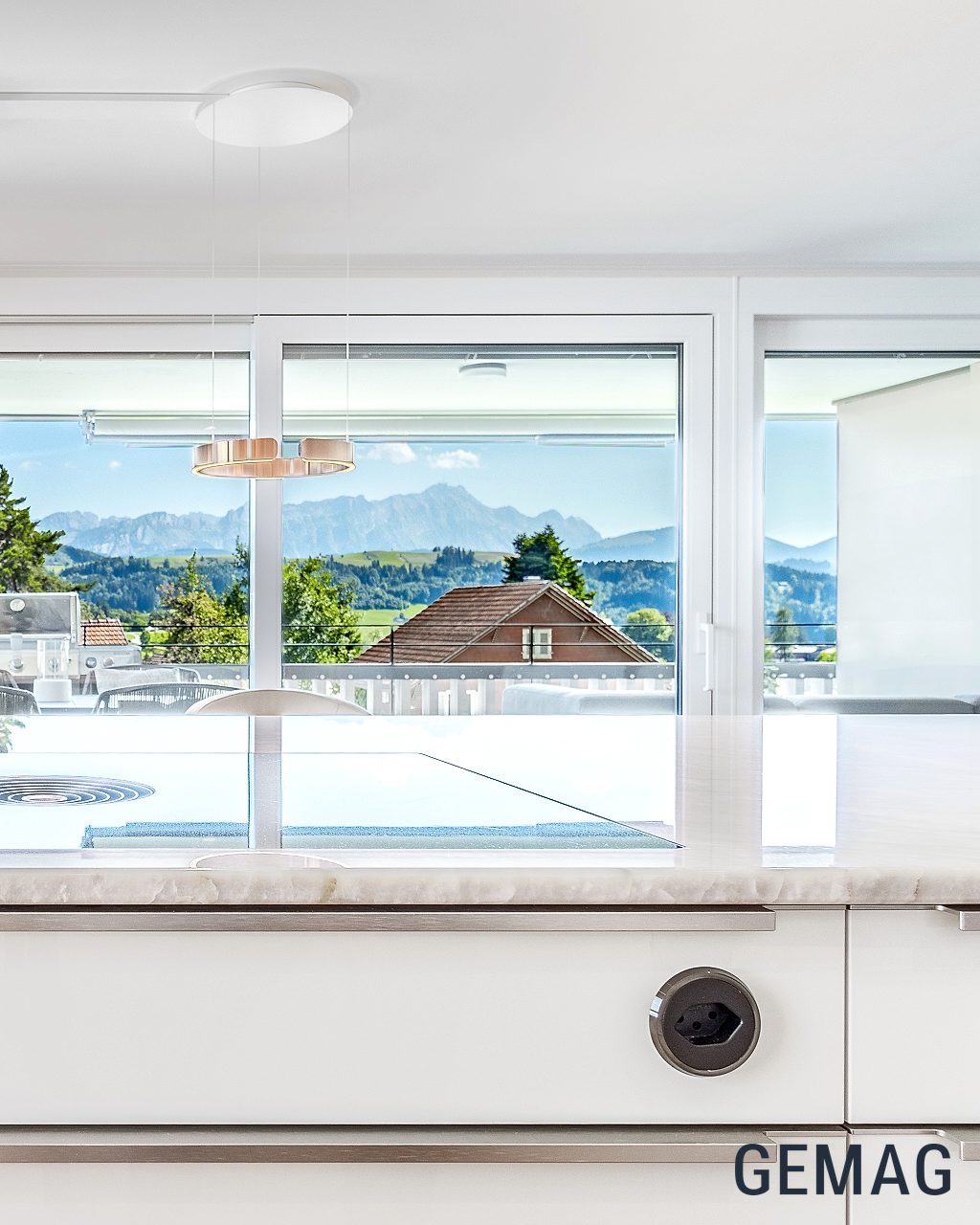 Kücheninsel mit Steinarbeitsplatte und Induktionskochfeld, im Hintergrund Schiebetür mit Blick auf Terrasse und Bergpanorama.