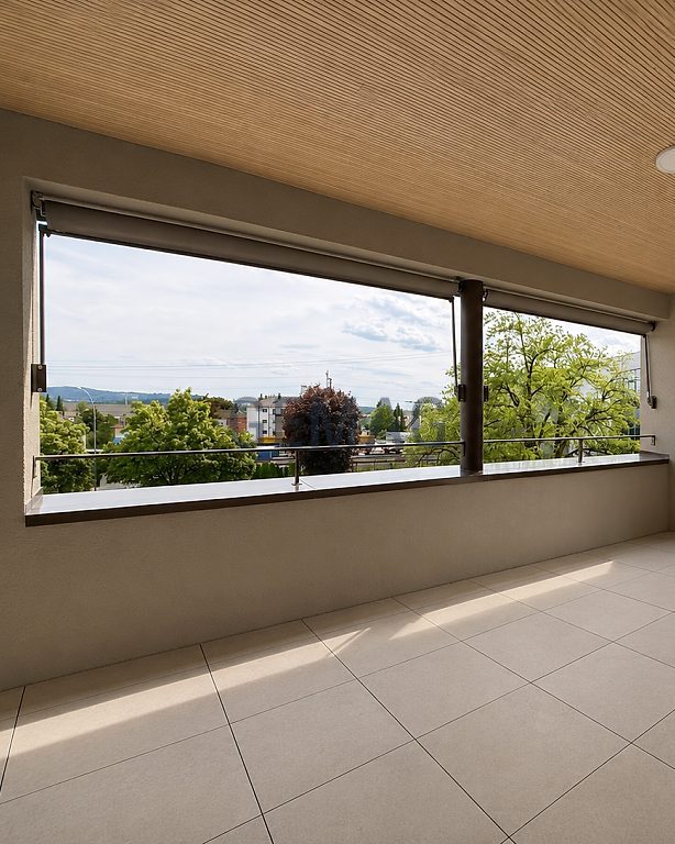 Überdachte Terrasse mit hellem Fliesenboden, Panoramafenstern und dekorativem Metallgeländer, Blick ins Grüne.