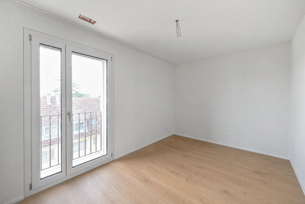 Leeres Zimmer mit Parkettboden, weissen Wänden und zweiflügeliger Balkontür, Blick in die Ecke.