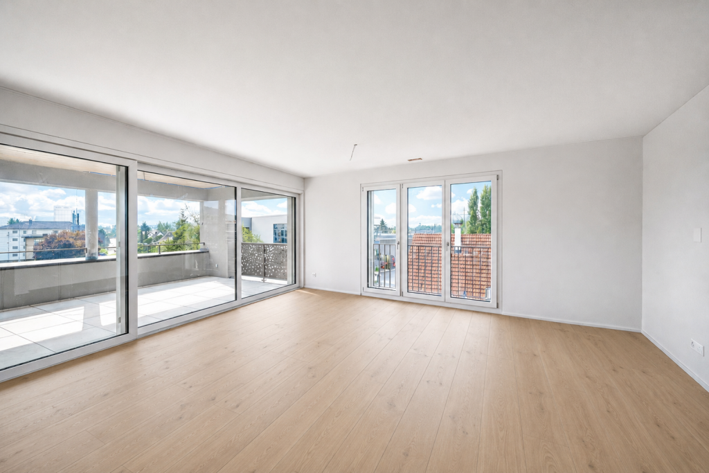 Leeres Wohnzimmer mit Holzboden, grosser Schiebetürfront zum Balkon und zusätzlichem Fenstertürelement, Frontalansicht.
