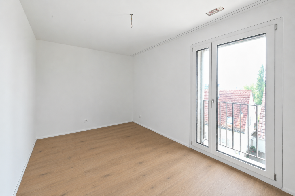 Leeres Zimmer mit Holzboden, weissen Wänden und zweiflügeliger Balkontür, Blick auf umliegende Dächer.