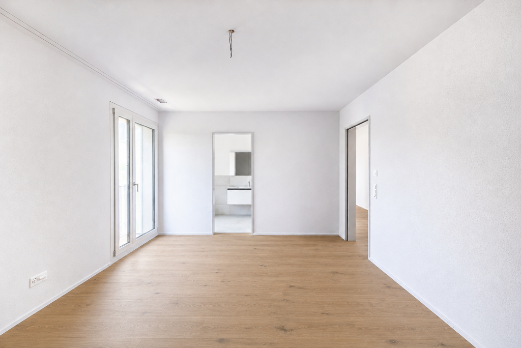 Leeres Zimmer mit Holzboden, weissen Wänden, Fenstertür und Blick in ein angrenzendes Bad.