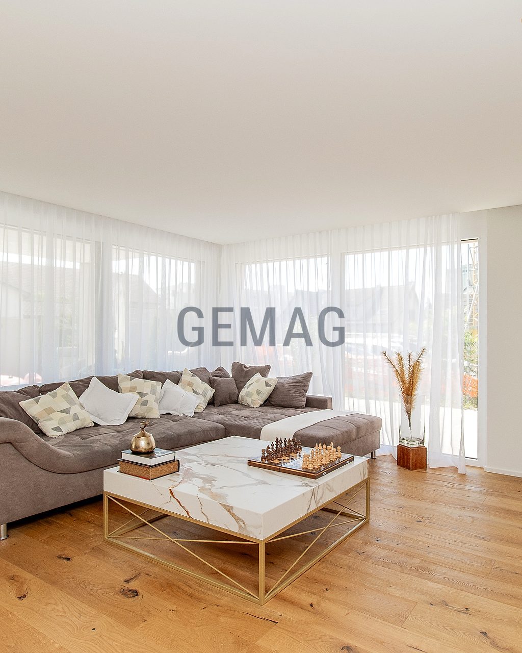 Wohnzimmer mit grossem Ecksofa, Marmor-Couchtisch, Parkettboden und raumhohen Fenstern mit weissen Vorhängen.