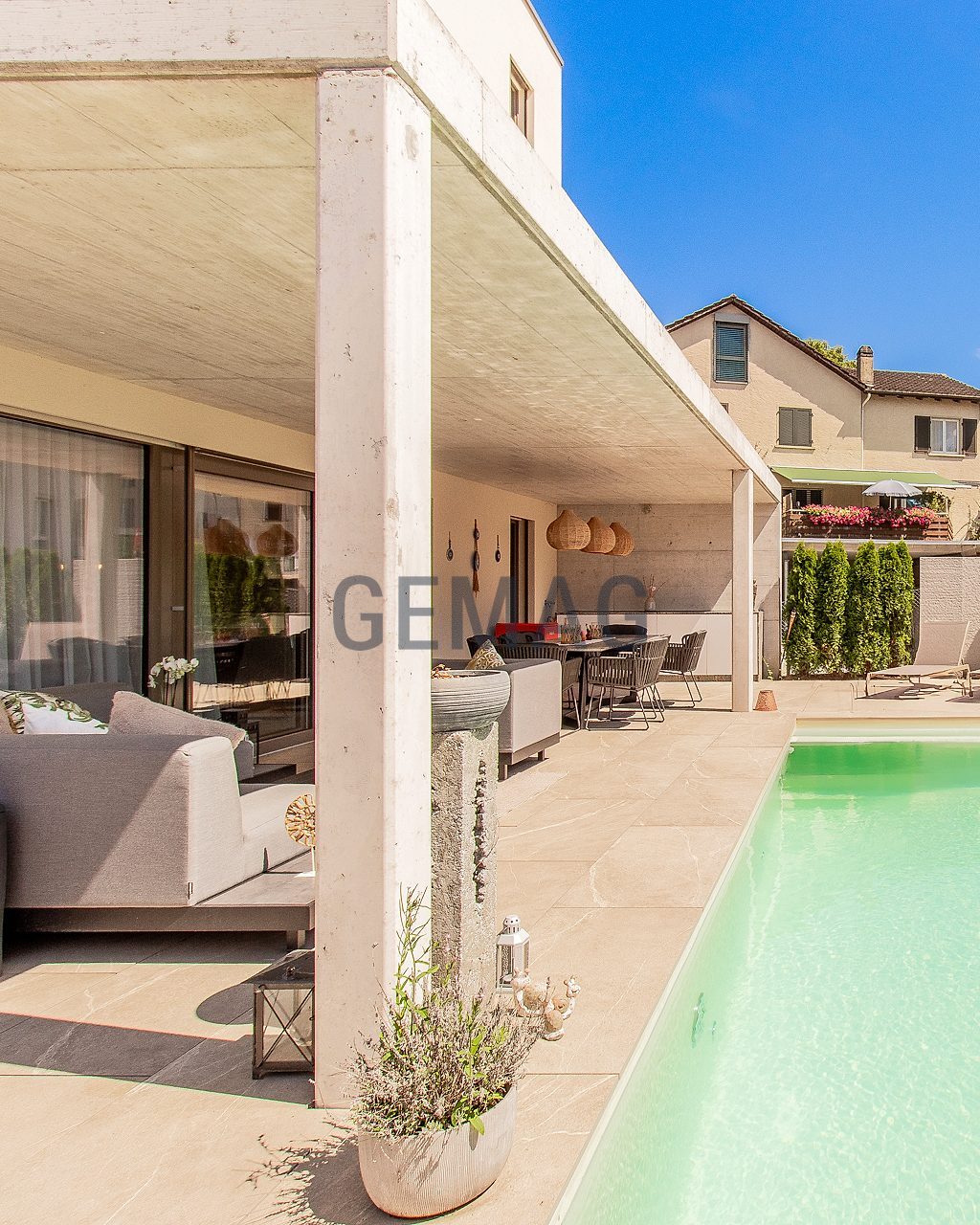 Überdachte Terrasse mit Aussensofa und Esstisch neben einem Swimmingpool, flankiert von Thujen, bei blauem Himmel.