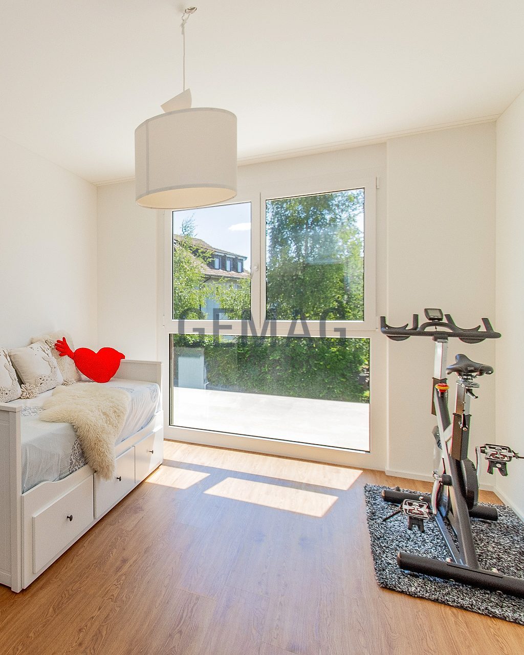 Zimmer mit Einzelbett, Holzboden und grossem Fenster mit Gartenaussicht; rechts ein Ergometer.