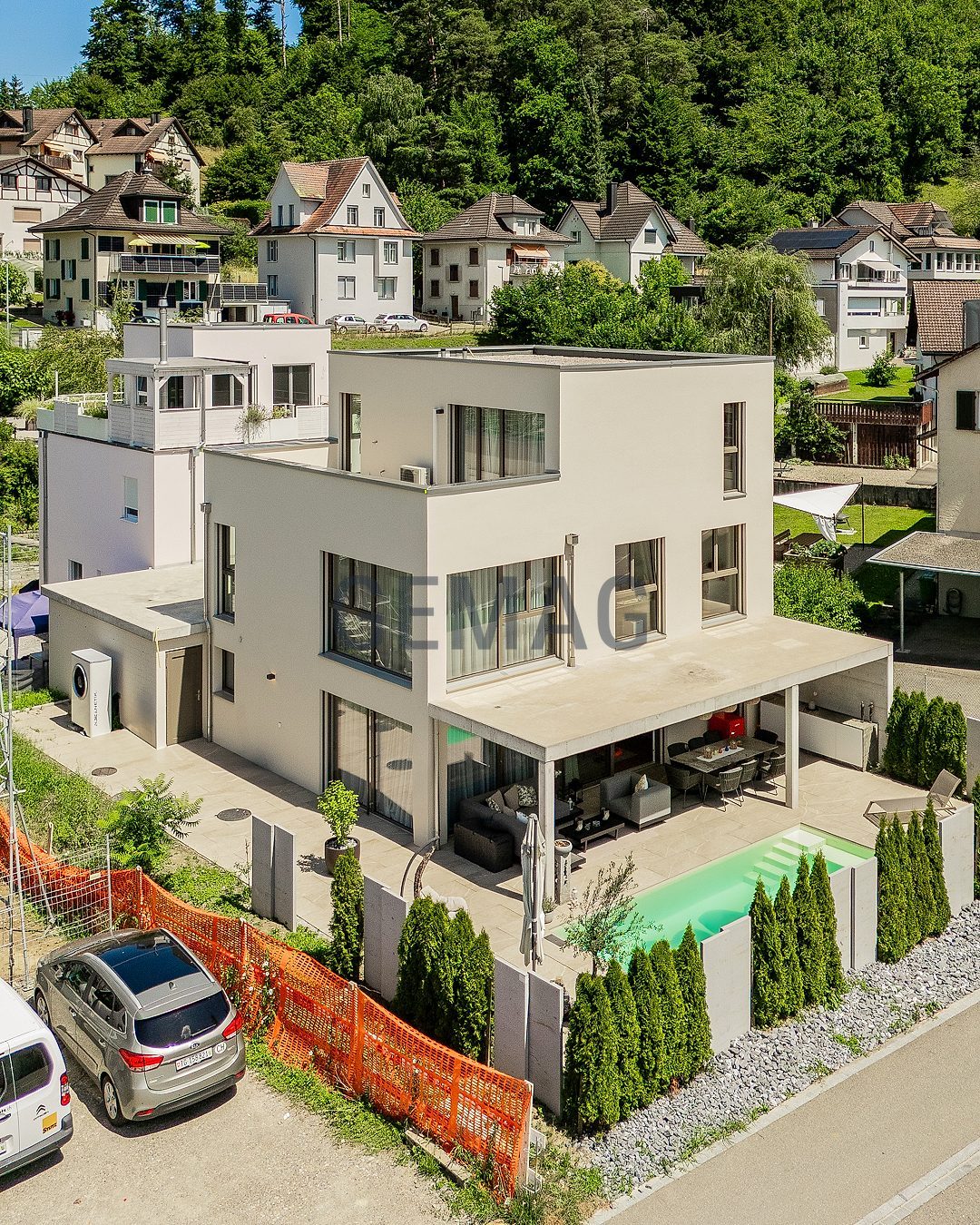 Drohnenaufnahme eines modernen Einfamilienhauses mit überdachter Terrasse, Pool und Thujahecke vor begrüntem Hügel.