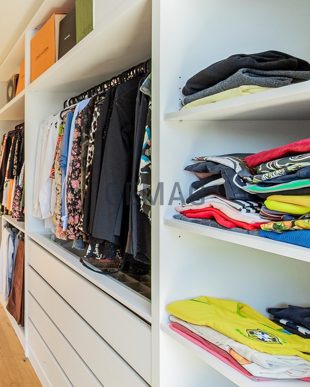 Langer begehbarer Kleiderschrank mit weissen Einbauregalen, Kleiderstangen und Schubladen, Holzboden, Fenster am Ende.