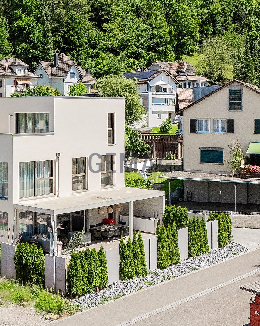 Drohnenaufnahme eines modernen Kubus-Einfamilienhauses mit überdachter Terrasse, Thujen-Hecke und umliegender Wohnbebauung vor Waldhang.