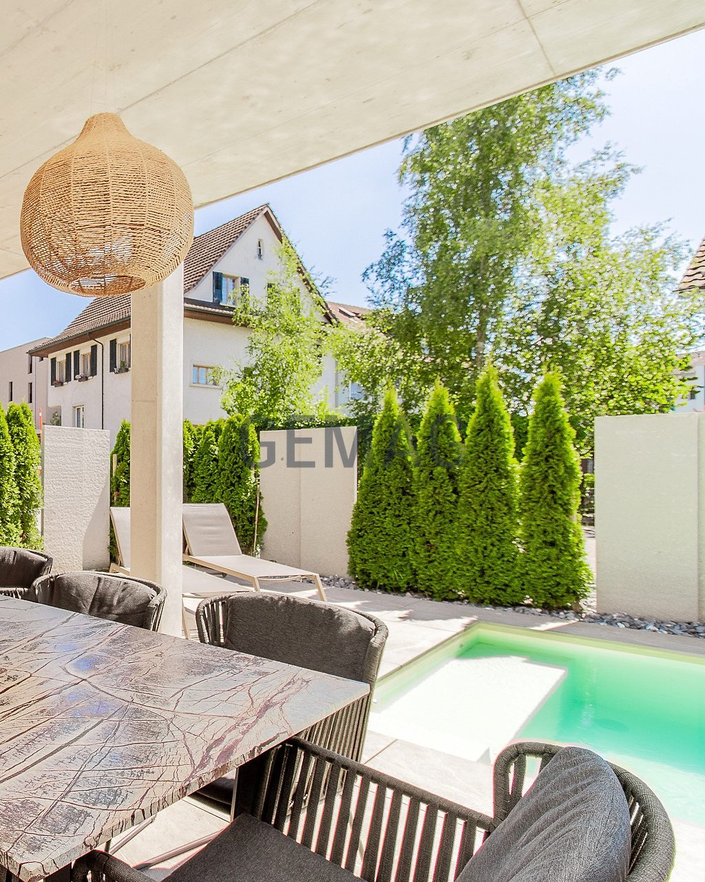 Überdachte Aussenlounge mit grossem Esstisch, Rattanleuchten und Blick auf den Pool und Thujenhecke.