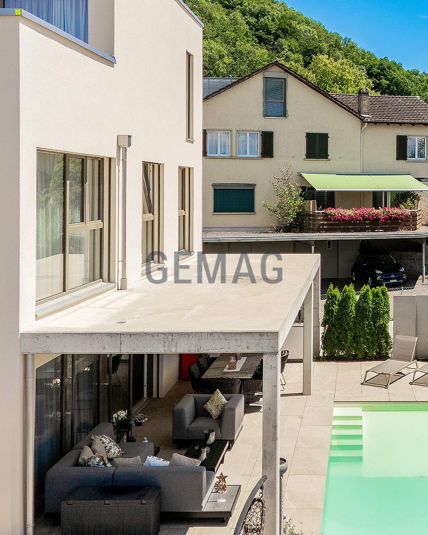 Luftaufnahme der Hausrückseite mit überdachter Terrasse, Pool und Liegebereich, dahinter Dorfstrasse und begrünter Hügel.