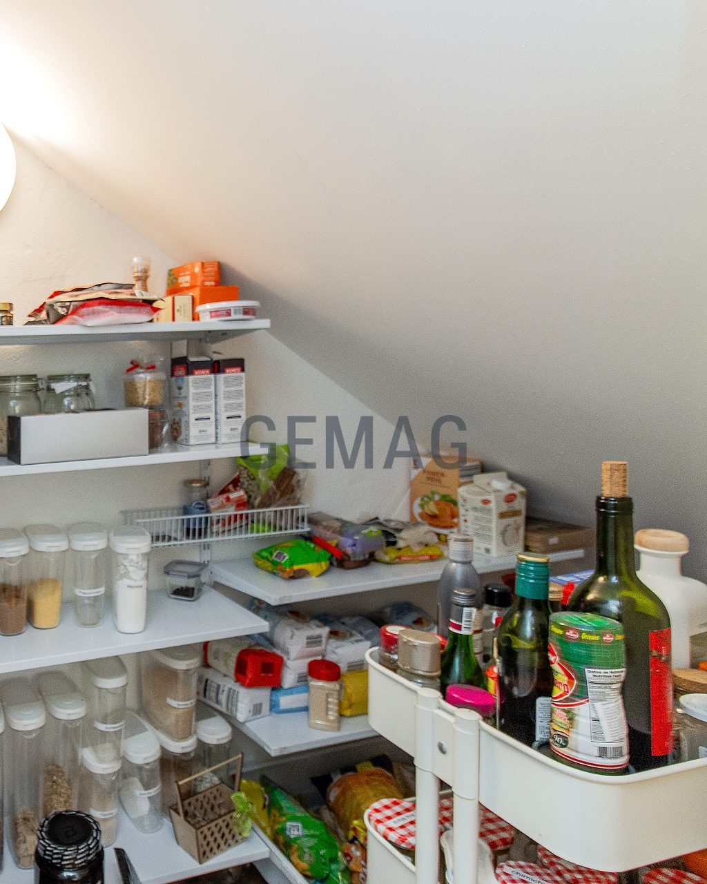 Vorratsraum unter einer Treppe mit weissen Wandregalen, gefüllt mit Lebensmitteln und Vorratsbehältern.