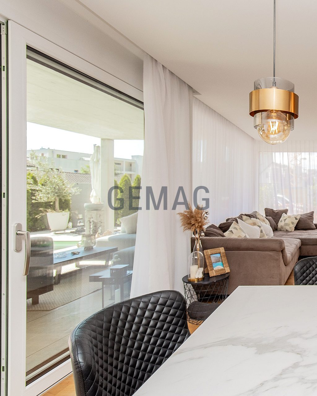 Offener Wohn-Essbereich mit Marmoroptik-Tisch, schwarzen Stühlen und goldenen Pendelleuchten, Blick zum Sofa und Terrassenzugang.