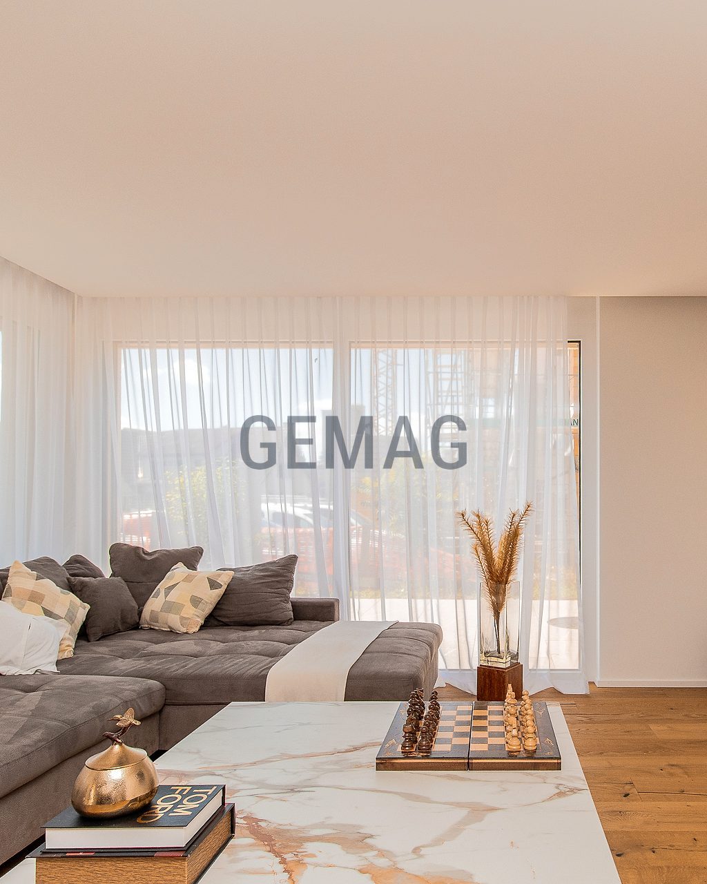 Wohnzimmer mit grossem Ecksofa, Marmor-Couchtisch, Parkettboden und bodentiefen Fenstern mit Gardinenzug.