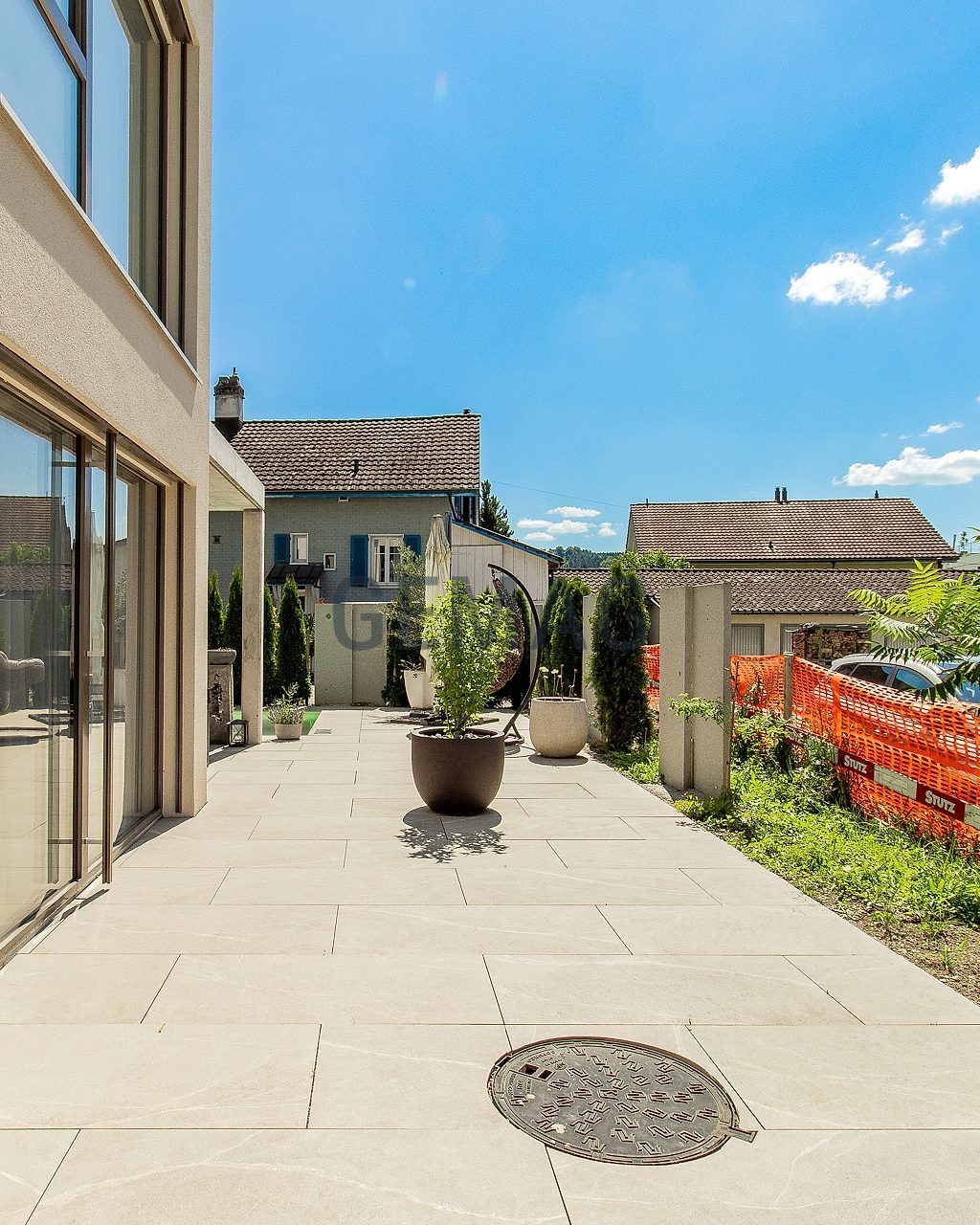 Geflieste Terrasse entlang der Hausfassade mit Schiebetüren, Kübelpflanzen und Blick auf umliegende Gebäude.