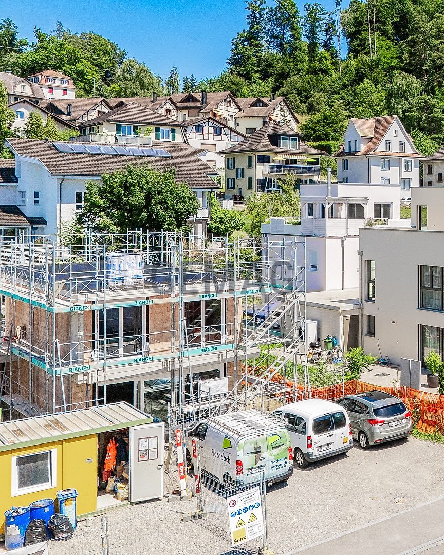 Luftaufnahme eines weissen Einfamilienhauses mit Flachdach, eingebettet in eine gewachsene Wohnsiedlung mit Waldhintergrund.