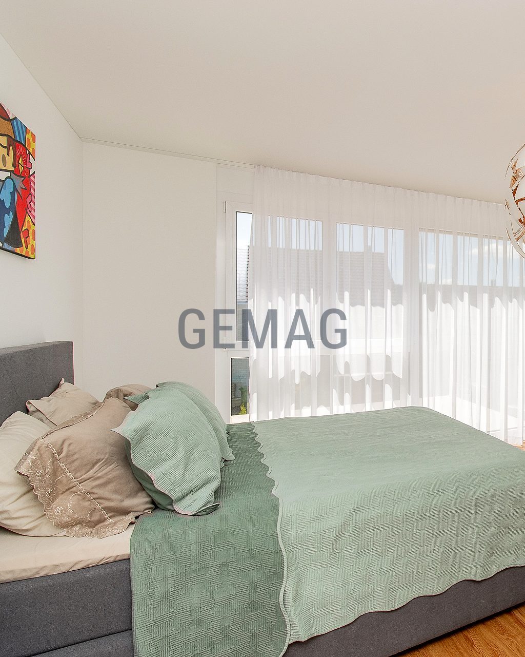 Schlafzimmer mit Doppelbett, Holzboden und grosser Fensterfront mit transparenten Vorhängen, Blick von der Seite.