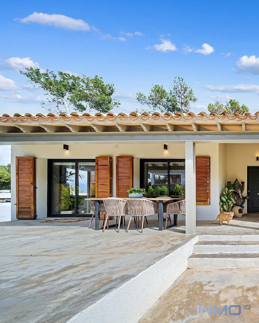 Frontansicht eines weißen Gebäudes mit Tonziegeldach, überdachter Terrasse mit Esstisch und Holzläden an den Fenstern.